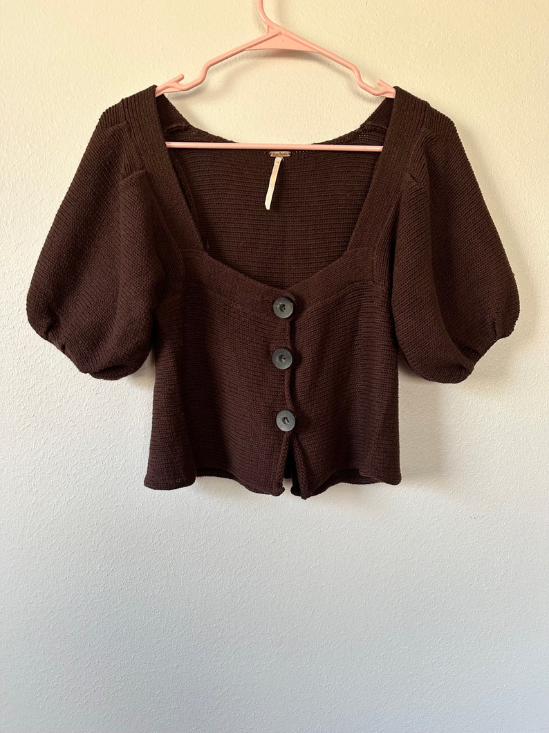 May include: A brown knitted cardigan with a square neckline and puff sleeves. It has a button closure and a cropped length.