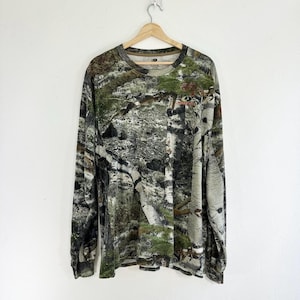 May include: A long-sleeved shirt with a camouflage pattern featuring shades of green, brown, and white. The shirt has a crew neck and a relaxed fit.