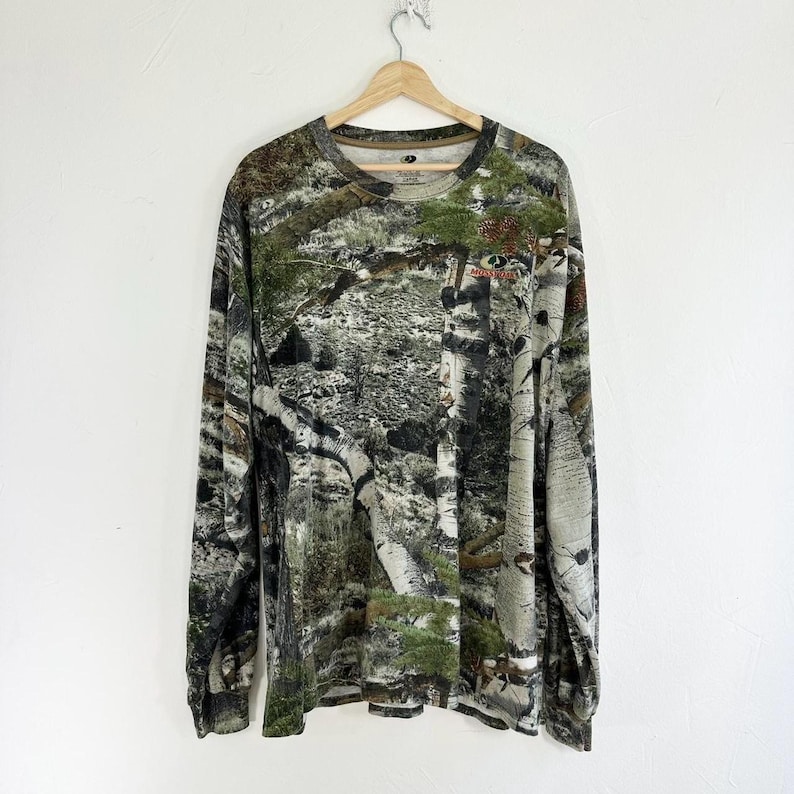 May include: A long-sleeved shirt with a camouflage pattern featuring shades of green, brown, and white. The shirt has a crew neck and a relaxed fit.