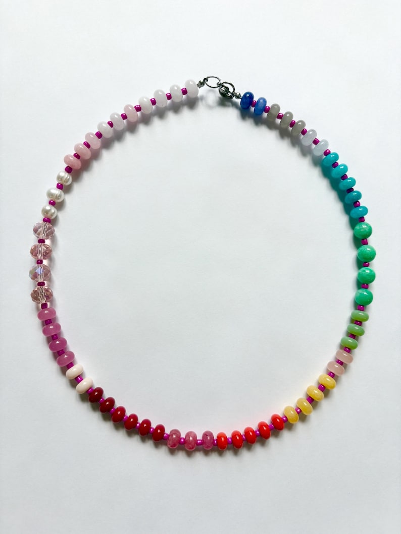 May include: A colorful beaded necklace with a variety of round beads in a rainbow of colors, including pink, red, yellow, green, and blue. The beads are strung on a pink cord and secured with a silver clasp. A unique piece of jewelry.