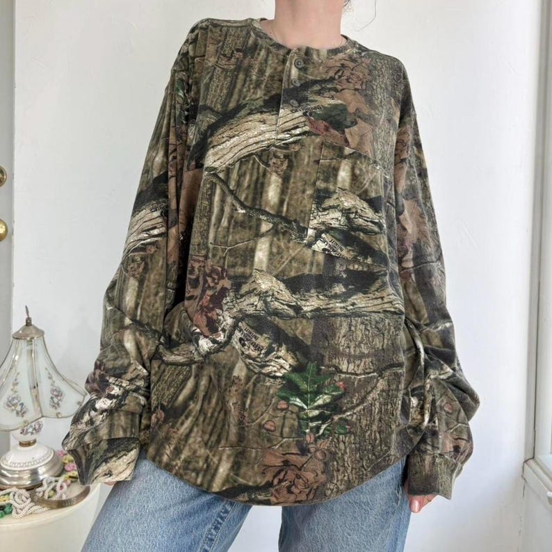 K&ouml;nnte beinhalten: Lang&auml;rmeliges Camouflage-Shirt mit Knopfleiste. Das Shirt hat ein Waldmuster in Braun-, Gr&uuml;n- und Schwarzt&ouml;nen. Das Shirt wird mit hellblauen Jeans kombiniert.
