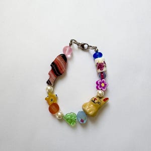 May include: A colorful beaded bracelet featuring a variety of decorative beads. The bracelet includes a pink bead, a striped face bead, a yellow cat bead, and a flower bead. The bracelet is secured with a silver clasp.