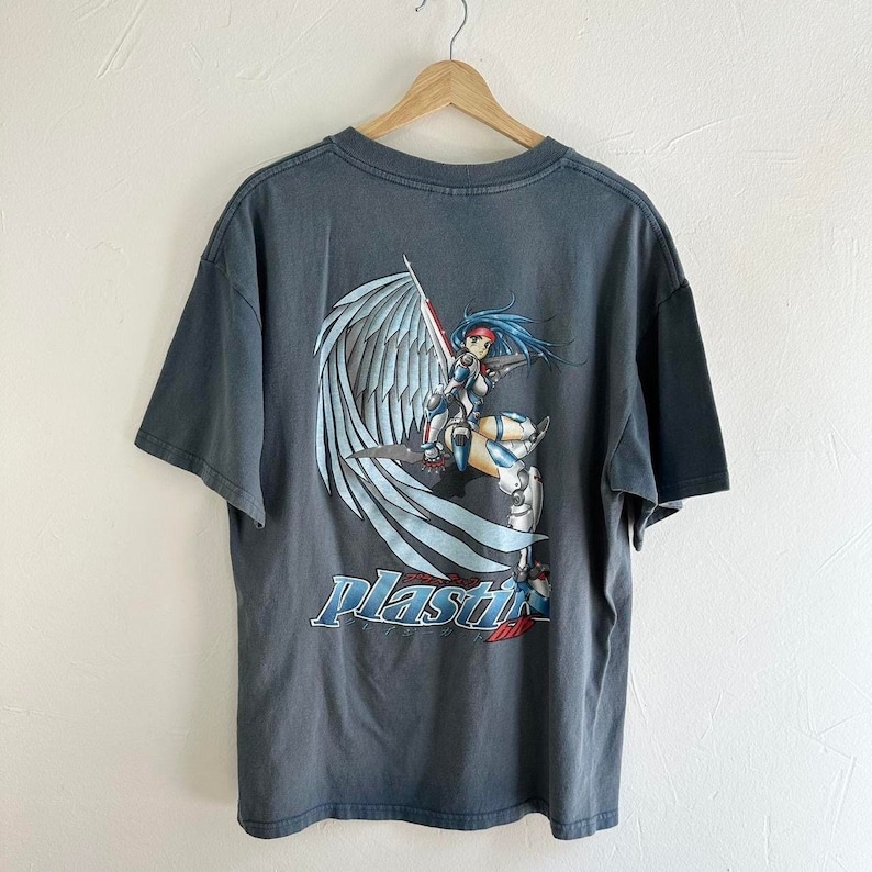 May include: A dark gray t-shirt with a graphic print of an anime-style woman with wings and a sword. The text "Plastic" is printed below the graphic.