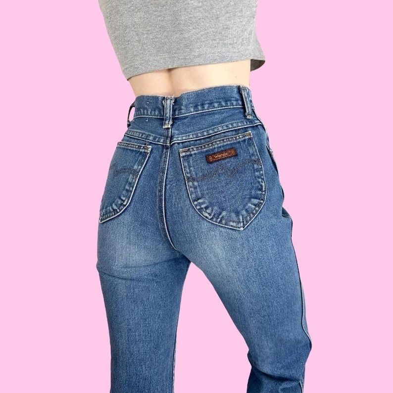 Puede incluir: Un par de pantalones vaqueros azules de cintura alta con un parche de cuero marr&oacute;n en el bolsillo trasero que dice "Wrangler".