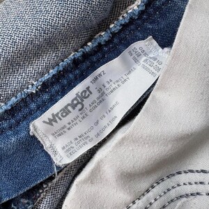 May include: Close-up of a Wrangler jeans label. The label is white with black text, displaying the brand name, size 33 x 34, and care instructions. The jeans are a medium-wash blue denim, with visible stitching and a textured appearance.