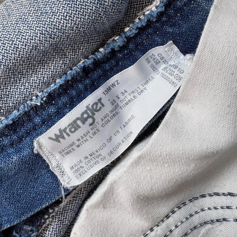 May include: Close-up of a Wrangler jeans label. The label is white with black text, displaying the brand name, size 33 x 34, and care instructions. The jeans are a medium-wash blue denim, with visible stitching and a textured appearance.