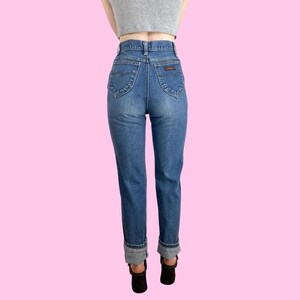 Puede incluir: Un par de pantalones vaqueros azules de cintura alta con dobladillo vuelto. Los vaqueros tienen un corte recto cl&aacute;sico y un lavado claro.
