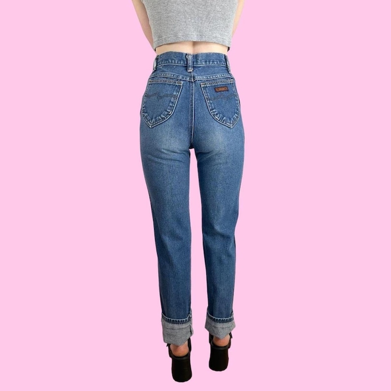 Puede incluir: Un par de pantalones vaqueros azules de cintura alta con dobladillo vuelto. Los vaqueros tienen un corte recto cl&aacute;sico y un lavado claro.