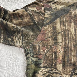 May include: Close-up of a camouflage pattern fabric. The fabric features a blend of brown, green, and tan hues, creating a woodland camouflage design. The word "BREAKUP" is visible on the fabric. The fabric is likely part of a garment.
