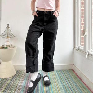 May include: Black denim jeans with large folded cuffs. The jeans have a classic five-pocket design and a button and zip closure. The model is wearing white socks and black Mary Jane shoes. The background includes a striped rug and a white wall.