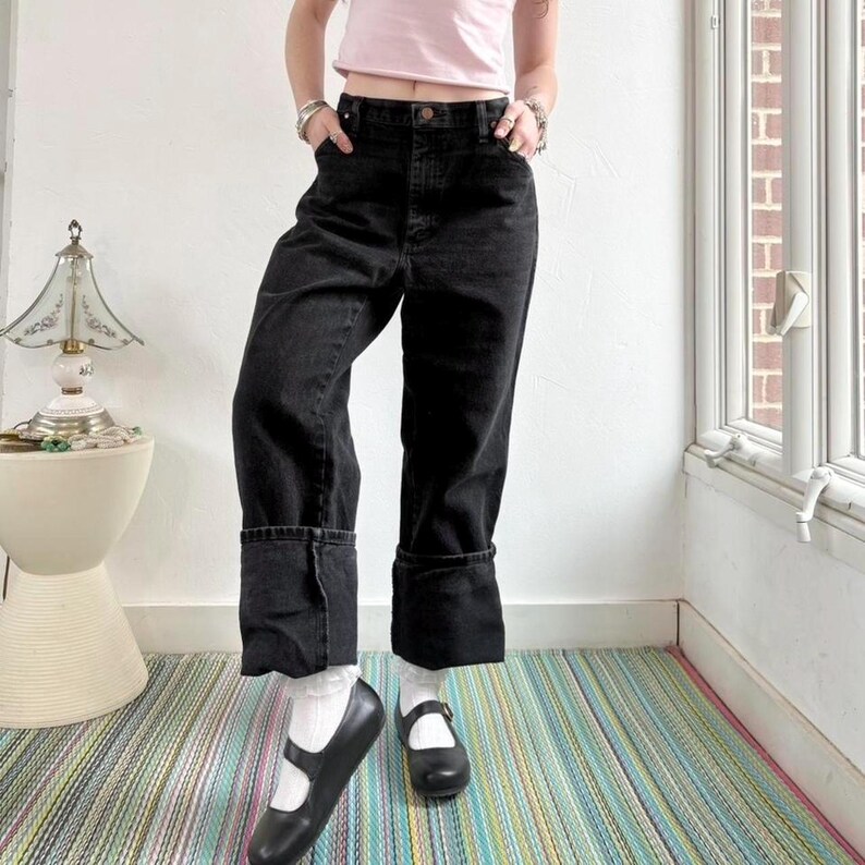 May include: Black denim jeans with large folded cuffs. The jeans have a classic five-pocket design and a button and zip closure. The model is wearing white socks and black Mary Jane shoes. The background includes a striped rug and a white wall.