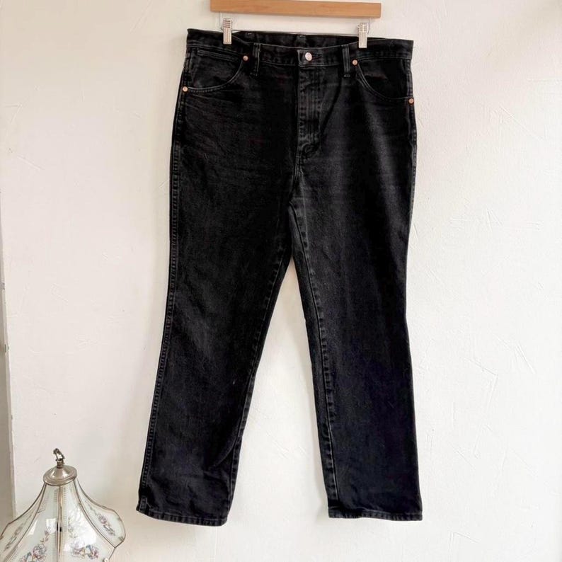 May include: Black denim jeans hanging on a wooden clothes hanger. The jeans have a classic five-pocket design with copper rivets and a button closure. The fabric is a dark wash, with subtle fading. The jeans are a straight-leg style.