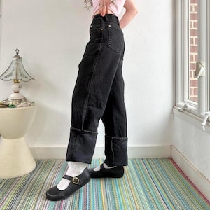 May include: Black denim jeans with a cuffed hem. The jeans are worn with black Mary Jane shoes with a gold buckle. The person is standing on a striped rug.