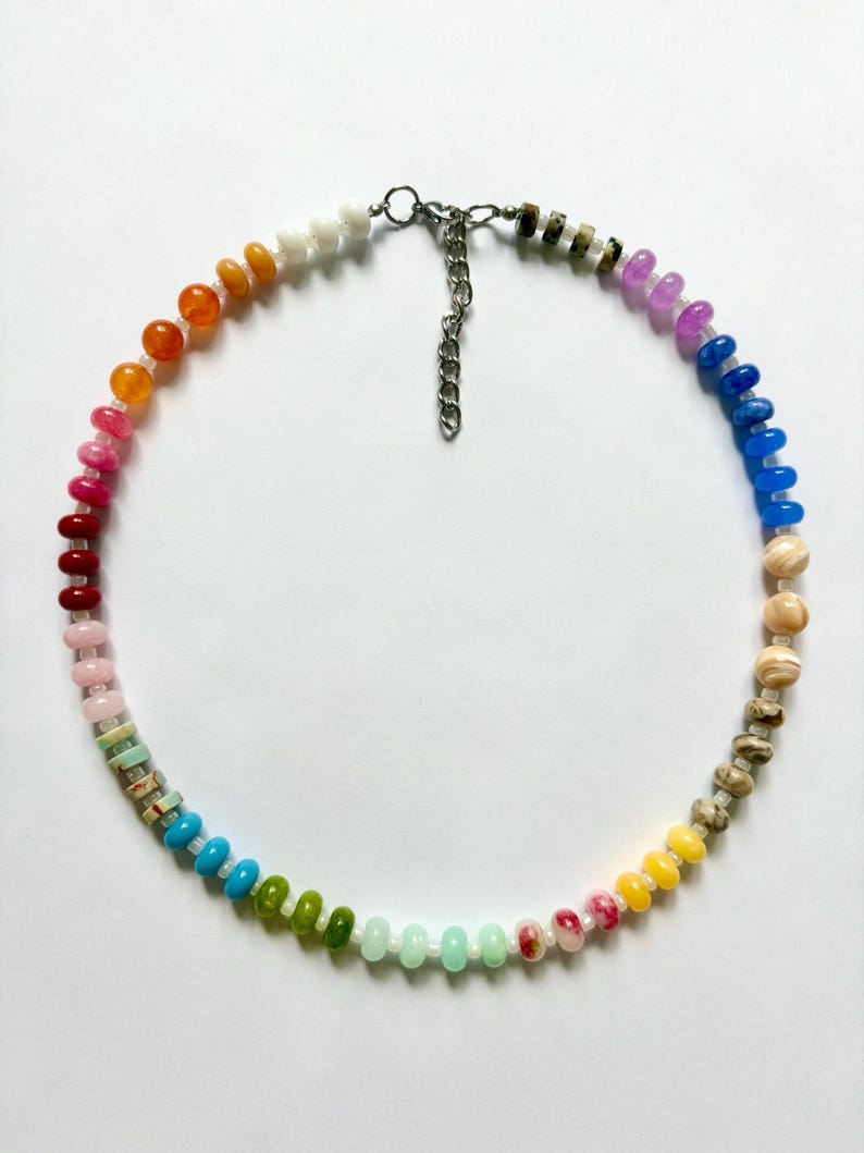 May include: A colorful beaded necklace featuring a rainbow of hues. The necklace showcases a variety of small, round beads in shades of red, orange, yellow, green, blue, and purple. The necklace has a silver-toned chain and clasp.