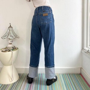 May include: Dark wash denim jeans with gray cuffs. The jeans have a high waist and a brown leather patch on the back pocket. The cuffs are approximately 3 inches in height. The person is wearing black shoes.