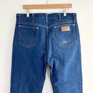 May include: A pair of blue denim jeans with a brown leather Wrangler label on the back pocket.