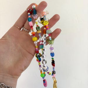 May include: A multi-coloured beaded lanyard featuring a variety of shapes including round, oval, and flower-shaped beads. The strand includes a yellow smiley face, a green butterfly, and white letter beads spelling out MRS BROOKE.