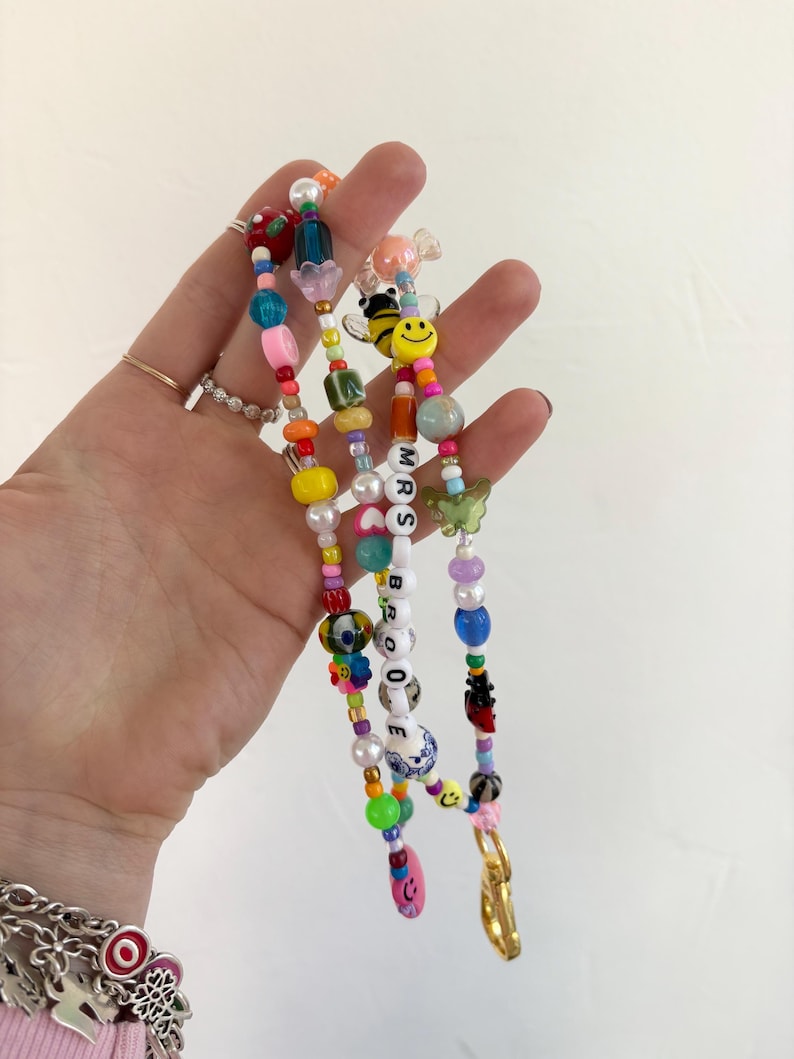 May include: A multi-coloured beaded lanyard featuring a variety of shapes including round, oval, and flower-shaped beads. The strand includes a yellow smiley face, a green butterfly, and white letter beads spelling out MRS BROOKE.