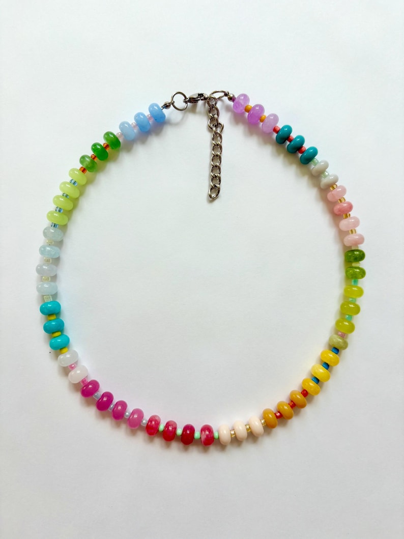 May include: A colorful beaded necklace with a rainbow gradient of colors. The necklace features small, round beads in shades of pink, red, yellow, green, blue, and purple. It has a silver-tone clasp and chain for adjustable length.
