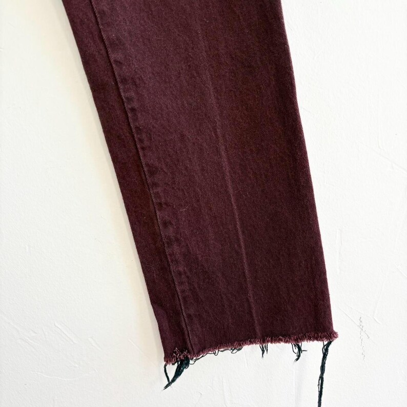 May include: Close-up of a pair of burgundy-colored jeans. The jeans have a straight leg cut and a frayed hem. The fabric appears to be denim. The background is a plain white surface.