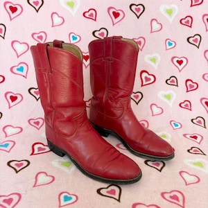May include: A pair of red leather cowboy boots with pointed toes. The boots are on a pink background with hearts.