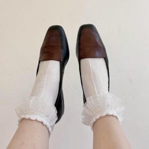 May include: A pair of black and brown leather loafers with square toes. The shoes are paired with white ribbed socks and white ruffled ankle cuffs. The image is set against a plain white background.