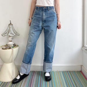 May include: Light blue denim jeans with a high waist and cuffed hems. The jeans have a classic five-pocket design and a faded, worn look. The cuffs are approximately 3 inches (7.6 cm) wide. The model is wearing black shoes.