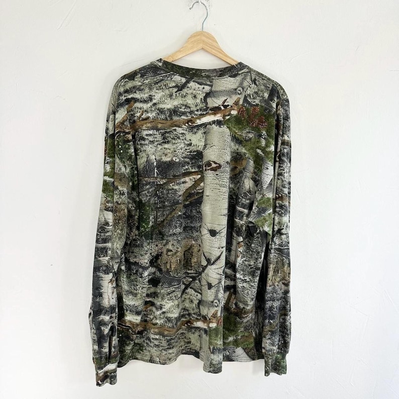 May include: A long-sleeved shirt with a camouflage pattern in shades of green, brown, and white. The shirt has a crew neck and a relaxed fit.