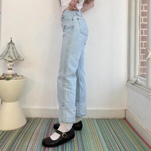 Op de afbeelding: Lichtblauwe denim jeans met omgeslagen zoom. De persoon draagt zwarte Mary Jane schoenen met een gouden gesp. Witte sokken. De jeans heeft een klassieke, rechte pijp.