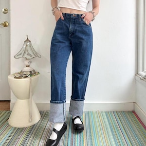 May include: Dark wash denim jeans with cuffed hems. The jeans have a classic five-pocket design and a straight leg fit. The model is wearing black Mary Jane shoes and white socks. The background includes a white wall, a small side table, and a colorful striped rug.