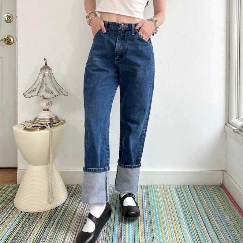 May include: Dark wash denim jeans with cuffed hems. The jeans have a classic five-pocket design and a straight leg fit. The model is wearing black Mary Jane shoes and white socks. The background includes a white wall, a small side table, and a colorful striped rug.