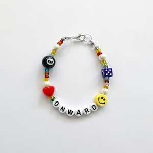 Puede incluir: Un brazalete de cuentas de colores con un dije de bola negra número 8, un dije de corazón rojo, un dije de dado azul, un dije de cara sonriente amarilla y cuentas de letras blancas que deletrean "ONWARD".