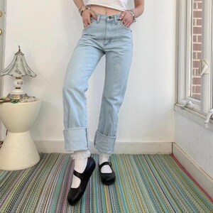 Op de afbeelding: Lichtblauwe spijkerbroek met omgeslagen zomen. De jeans hebben een klassiek ontwerp met vijf zakken. De persoon draagt zwarte Mary Jane-schoenen en witte sokken. De achtergrond omvat een gestreept tapijt en een witte muur.