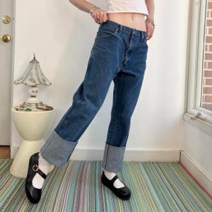 May include: Blue denim jeans with cuffed hems. The jeans are paired with black Mary Jane shoes. The outfit is on a striped rug. A white top is also visible.