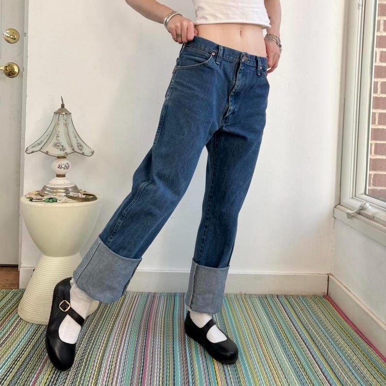 May include: Blue denim jeans with cuffed hems. The jeans are paired with black Mary Jane shoes. The outfit is on a striped rug. A white top is also visible.