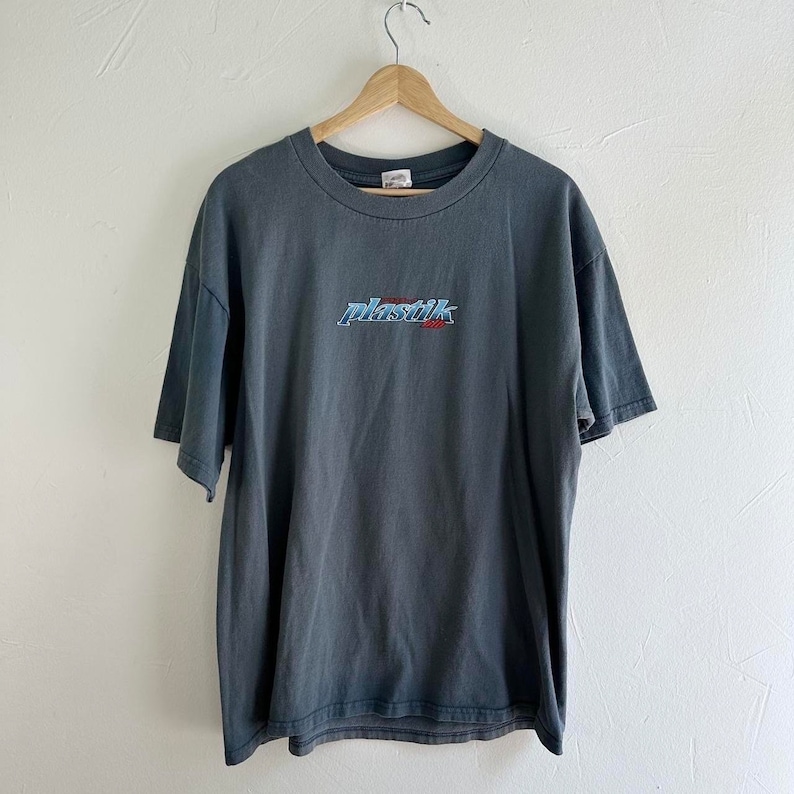 May include: A dark gray t-shirt with a blue and red embroidered logo that says "plastik" on the front.