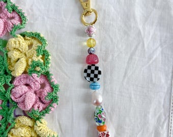 May include: A gold-colored keychain with a variety of colorful beads. The beads include a pink floral bead, a checkerboard bead, a pearl, and a floral glass bead. The keychain is attached to a gold-colored clasp.
