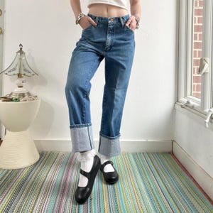 以下が含まれることがあります： 裾を折り返したブルーデニムジーンズ。ジーンズは、クラシックな5ポケットのデザインとボタンとジッパーの留め具を備えています。 モデルは黒いメリージェーンシューズを履いています。