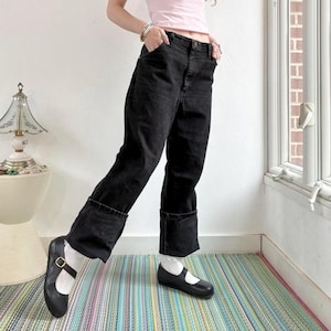 May include: Black denim jeans with turned-up cuffs, worn with black Mary Jane shoes and white socks. The jeans feature a five-pocket design. The shoes have a strap and buckle. The outfit is casual and fashionable.