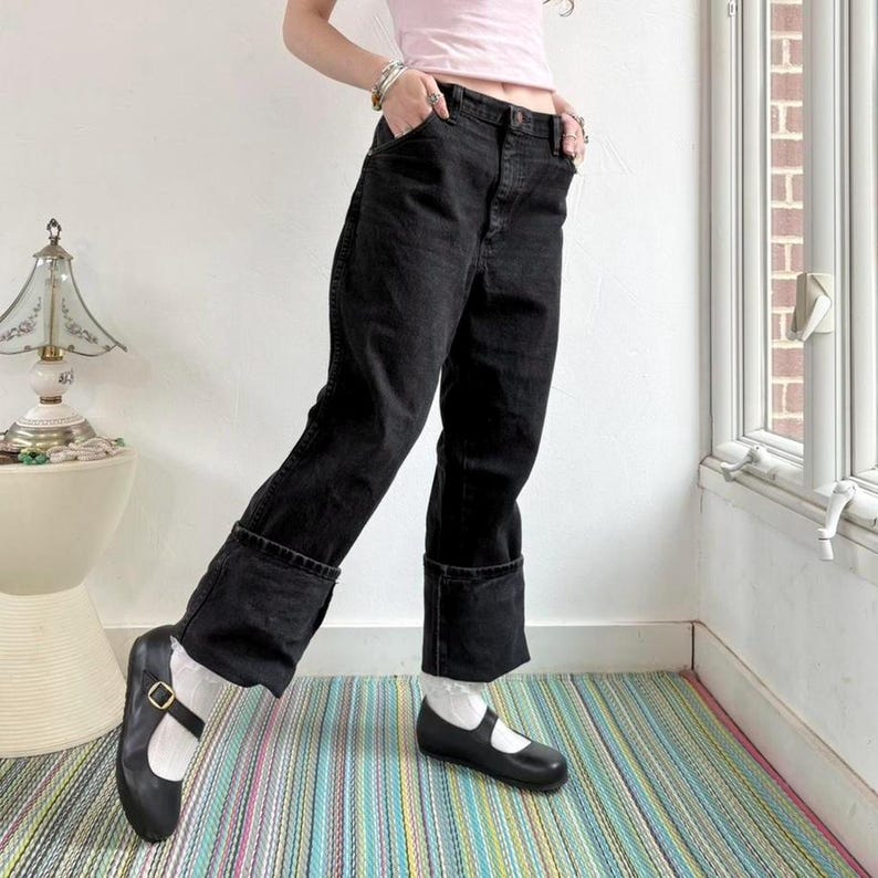 May include: Black denim jeans with turned-up cuffs, worn with black Mary Jane shoes and white socks. The jeans feature a five-pocket design. The shoes have a strap and buckle. The outfit is casual and fashionable.