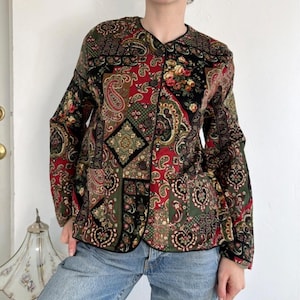 May include: A long-sleeved jacket featuring a patchwork design with a mix of red, black, green, and gold colors. The fabric displays intricate paisley patterns and floral motifs. The jacket has a round neckline and a front button closure.