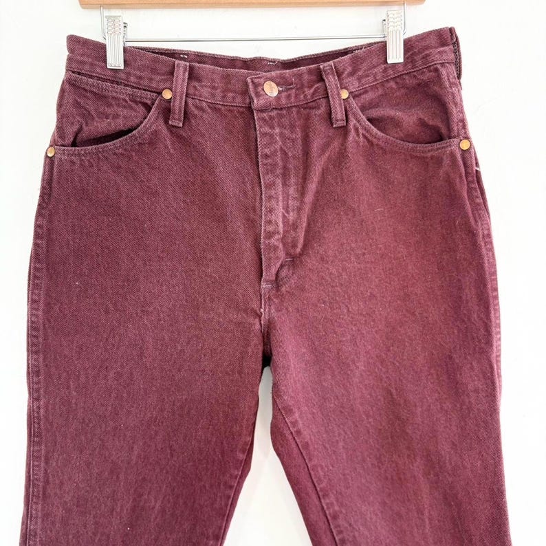 May include: Close-up of burgundy-colored denim jeans. The jeans are hanging on a wooden hanger. The jeans have a classic five-pocket design with copper-colored rivets and a button closure. The fabric appears to be slightly faded, giving it a vintage look.