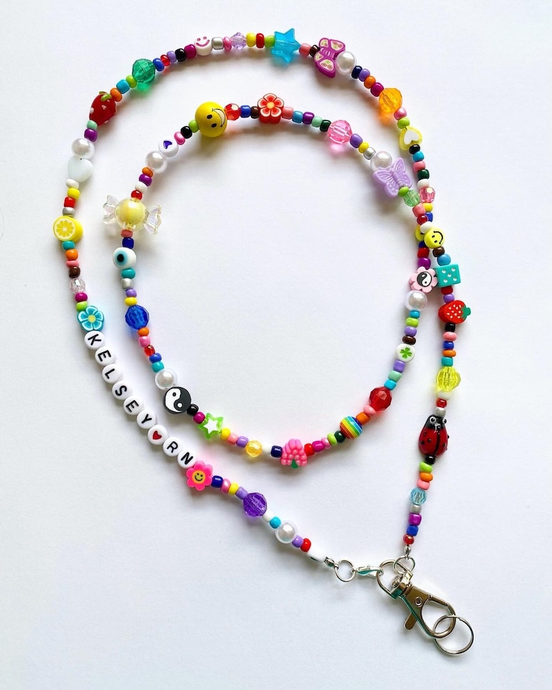 May include: A colorful beaded phone chain with a silver clasp. The chain features a variety of charms, including a smiley face, a butterfly, a flower, a star, a candy, an eye, a yin yang symbol, a ladybug, a strawberry, and a heart. The chain also includes the name "Kelsey" spelled out in white beads.