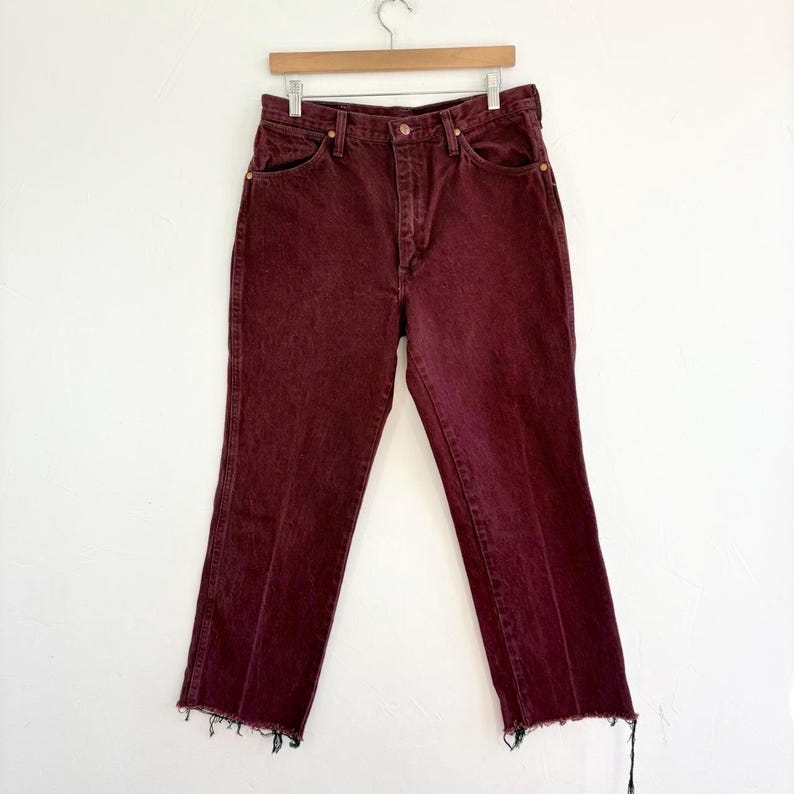 May include: Burgundy-colored denim jeans with a straight leg and a raw, frayed hem. The jeans are hanging on a wooden hanger against a white background. The jeans have classic five-pocket styling and a button and zipper closure.