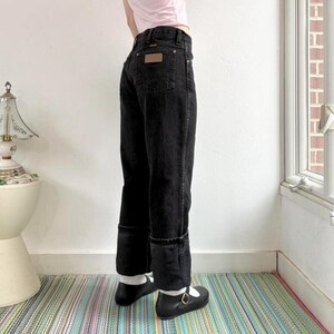 May include: Black denim jeans with a brown patch on the back pocket. The jeans have a straight leg and are cuffed at the ankles. The person is wearing black shoes with a gold buckle.
