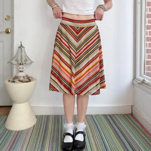 May include: A knee-length skirt featuring a chevron pattern with horizontal stripes in shades of orange, pink, yellow, black, and white. The skirt has a fitted waistband and a flared silhouette, constructed from a knit fabric with a visible texture.