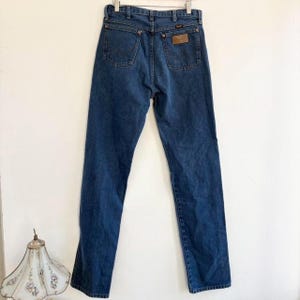 May include: Dark blue denim jeans with a classic five-pocket design. The jeans feature a brown leather patch on the back pocket with a brand logo. The jeans are hanging on a metal hanger against a white background.