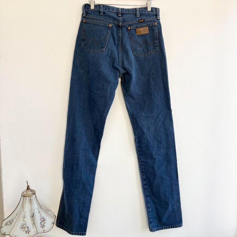 May include: Dark blue denim jeans with a classic five-pocket design. The jeans feature a brown leather patch on the back pocket with a brand logo. The jeans are hanging on a metal hanger against a white background.