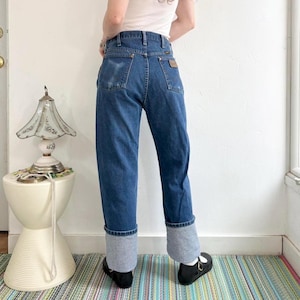 May include: Blue denim jeans with a light gray cuff. The jeans have a classic five-pocket design and a brown leather patch on the back. The jeans are worn with black shoes.