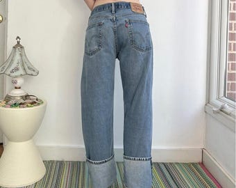 May include: Light blue denim jeans with a high-waisted fit, featuring wide legs and prominent folded cuffs at the hem. The back includes two pockets with arcuate stitching, a small red brand tab, and a tan leather patch on the waistband.